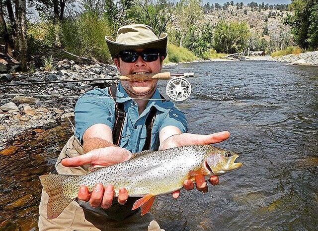 A man holds a fish out while a fishing pole in between his mouth.