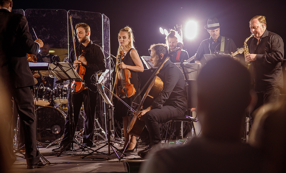 Musicians playing violin, violoncello and saxophone while conductor directing orchestra performance on the street at night